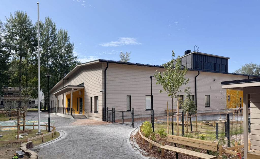 Päiväkodin leikkipiha, joka toteutettu mikrobiystävällisenä. Hirsirakennus, jossa on vaalea julkisivu.